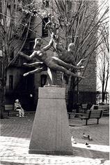 Black and white photograph of a sculpture in a park. The sculpture depicts several dancing figures leaping sideways through the air.