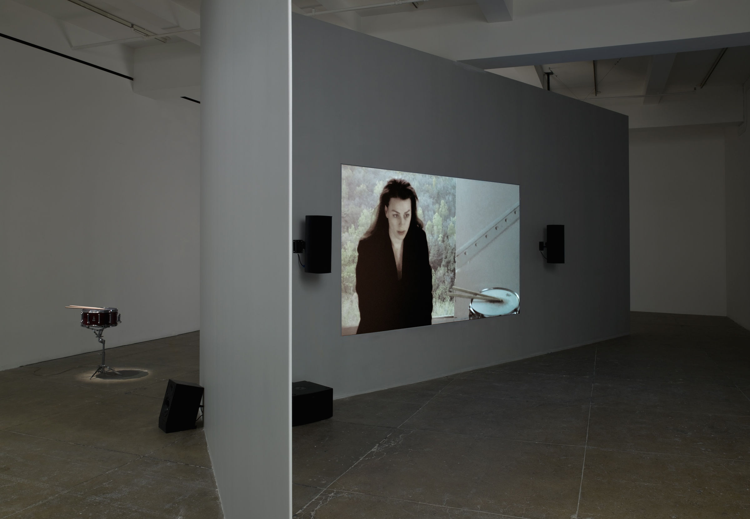 A projection of a woman is centered on a floating wall; 4 black speakers are visible around the screen and a snare kit sits behind the wall. 