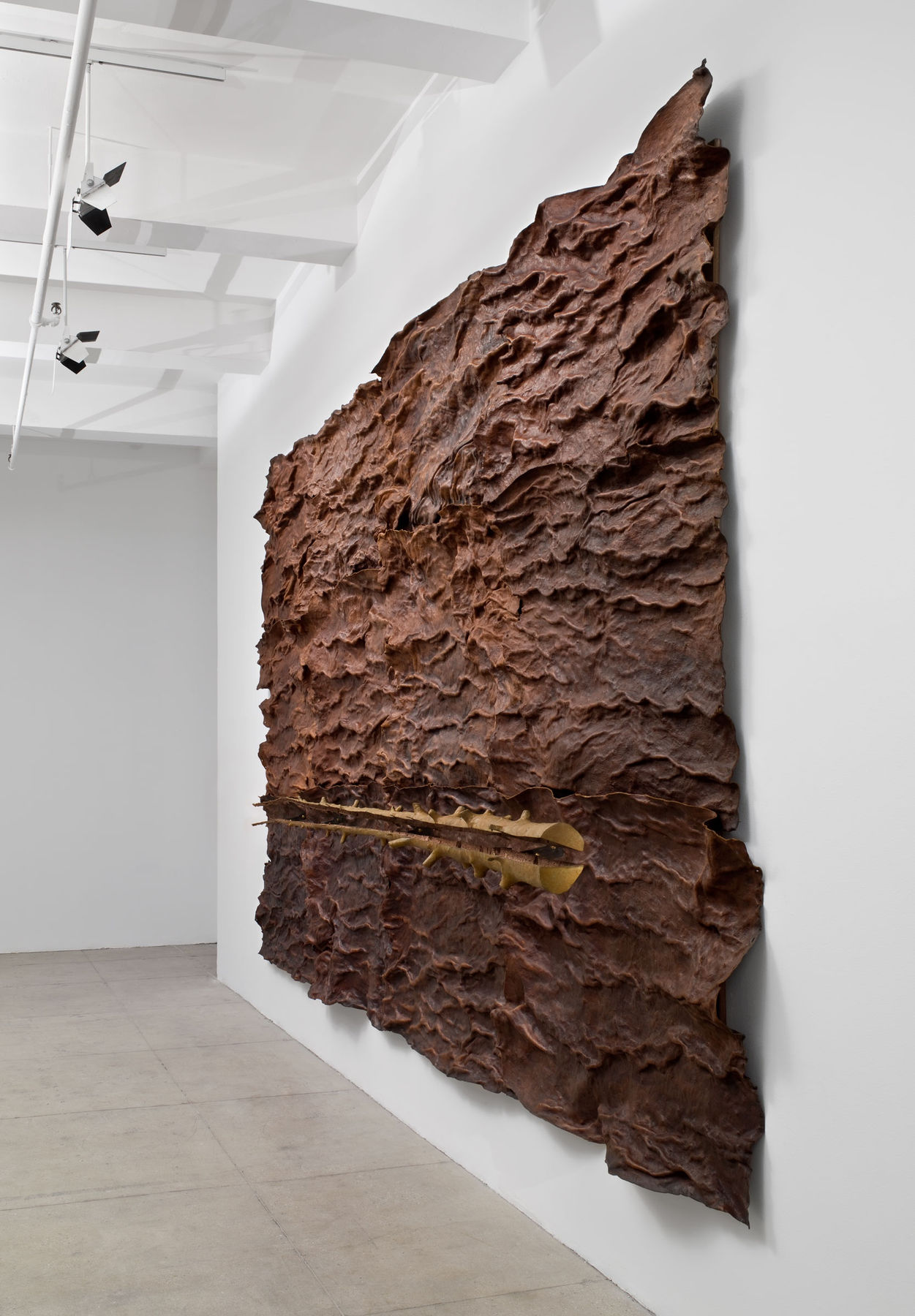 A large bark-like sculpture with a tree trunk running horizontally in front of it hangs on a white wall. 