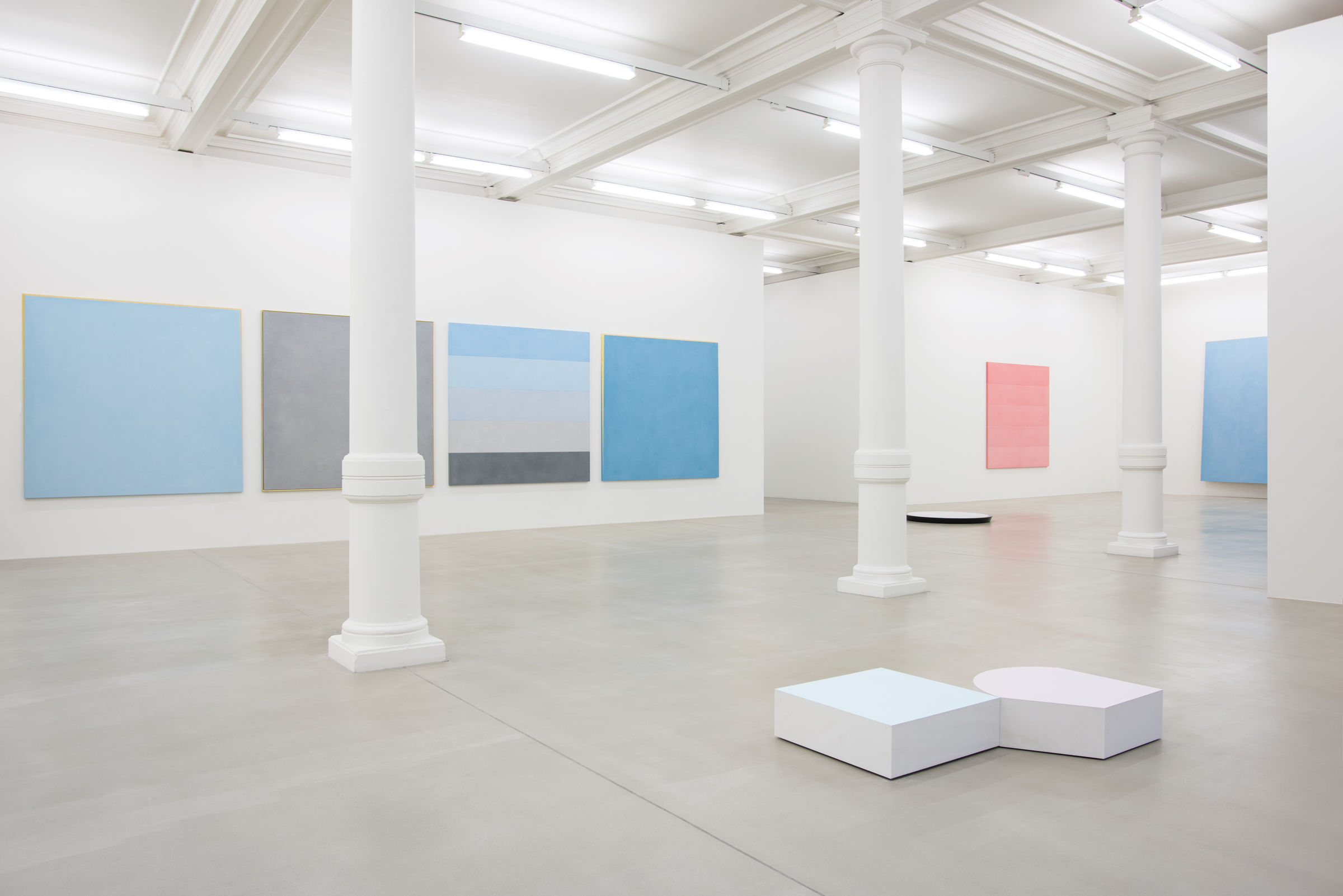 Various paintings of light shades (blue, grey, pink) line the walls of an all white space, with short white sculptures on the floor.