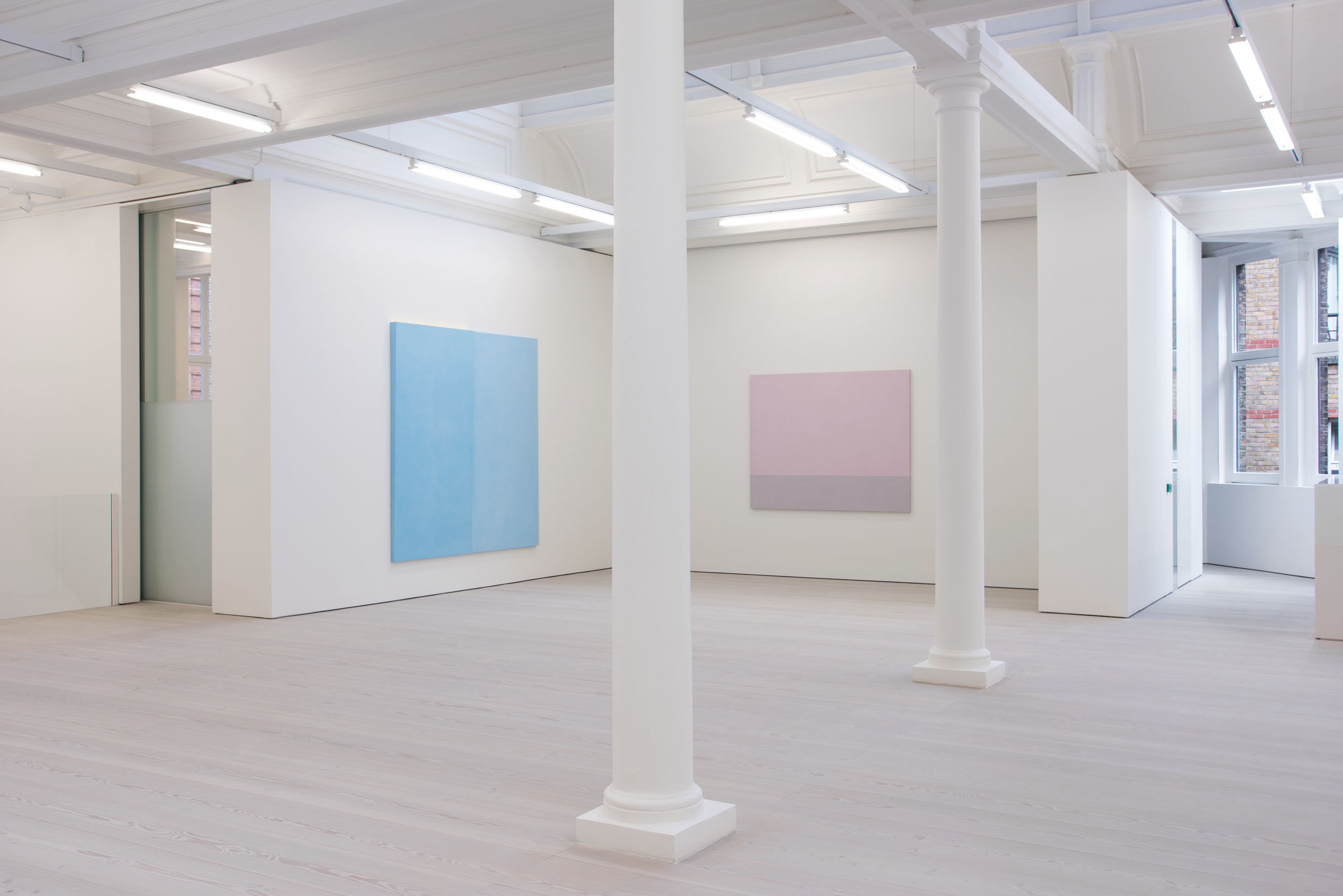 In a white gallery space with columns and a window to the left, two paintings hang: one light blue on the left wall, and one light pink straight ahead.