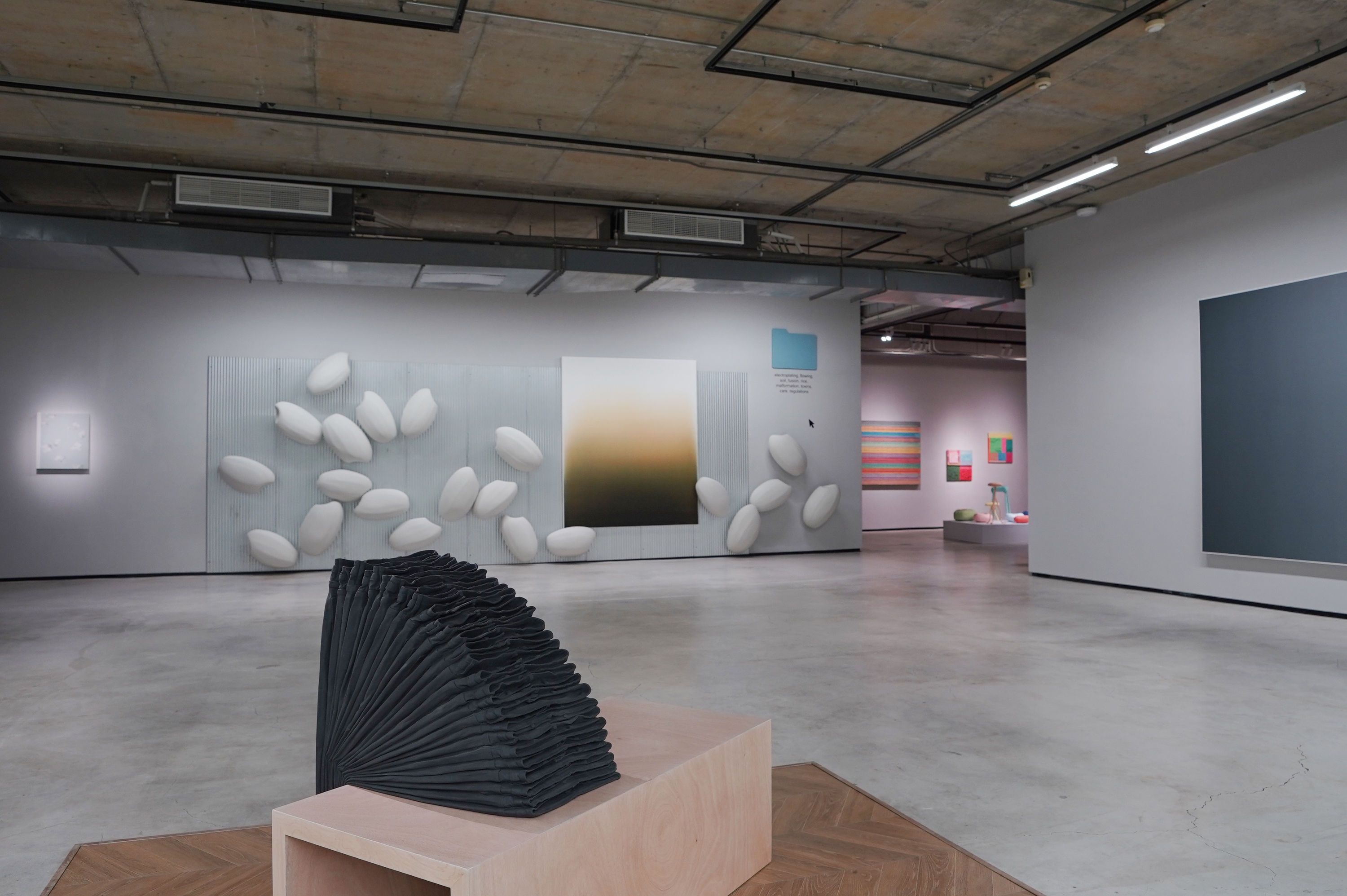 在超過三十年的創作時間,彌載映的圖像始終朝向一種抽象的表徵,長期以來,透過藝術行動的介入、日常空間的重塑,同時融入佛教法門的精神世界與社會實踐,屬熱帶氣候的鮮明色彩皆以顏料、肌理編碼進看似平靜和諧的繪畫、雕塑、裝置和參與式作品中。還有什麼比一句誓言換來一顆“石頭”更適合創造人群的聚集?原為參與式計畫的《Marking Stones》(2022),援引泰國佛寺中的結界石(Bai Sema)——它們界定出神聖的空間,也作為動土前的測量工具,如今轉化成猶如籃子、燈具、凳子造型的彩色紙漿雕塑,在展廳內部結界出可能的想像場域。 冷靜觀察——諸如日常環境中的污染、污漬都能調和進周育正的創作實踐。也許,這些信息可以重新轉譯成藝術語言,菜瓜布與抹布構建出繪畫行為,使「衛生」的概念在周育正的造型語彙下破題為《刷新、犧牲、新衛生、家居、 洗滌、周育正、 顏料、抹布、菜瓜布、畫布、 形象、繪畫集 #4》(2018)。在《電鍍金、流動、土壤、融合、稻米、畸形生長、毒素、關懷、法規》(2019),藝術家將常見於經濟型住宅的鍍鋁鋅板,結合大型米粒雕塑、漸層畫作、仿iOS系統的藍色文件夾、及滑鼠游標排列組合於牆面上。不僅源於環境污染的主題,作品從一連串的詞彙到圖像的轉譯,彷彿展示了當代網絡信息如何將對立的元素生成新意象,同時展開語言命名與視覺空間的複雜關係。