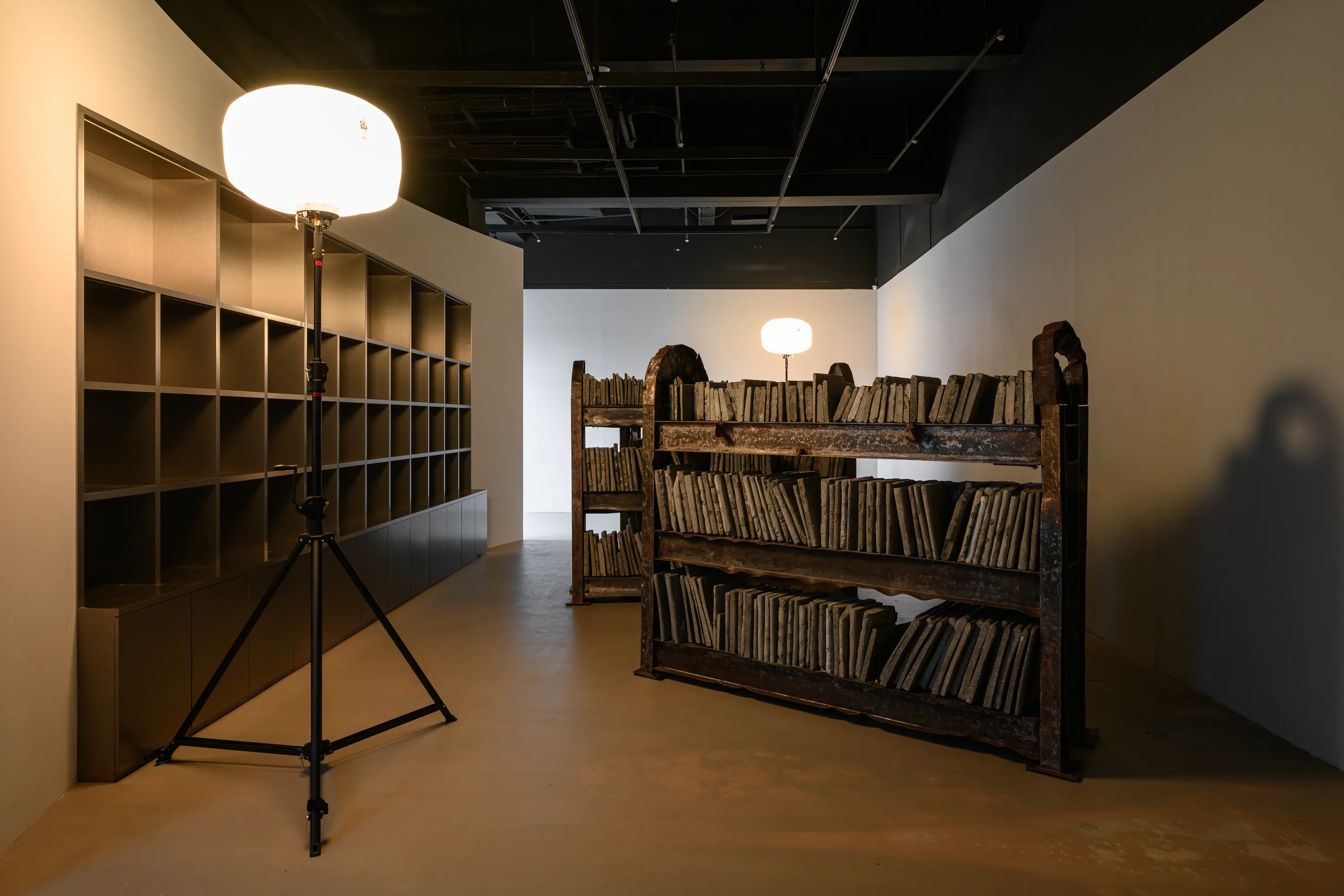 而在作品〈夜與靈魂〉中，藝術家將地震災後遺留的扭曲鋼筋與碎裂混凝土，轉化為書櫃與書本的雕塑結構，構築出一座承載記憶的微型圖書館。工作燈投射出的微光在金屬與碎片之間緩慢游移，使空間瀰漫著一層近乎潮濕的氣息，彷彿某段被壓覆於瓦礫之下的時間重新滲出表層。邱承宏曾提及，這件作品揉合了他在高壓狀態下反覆出現的夢魘意象，以及 0403 地震中倒塌建築所留下的斷壁殘垣。藝術家自廢棄場回收地震後遺留下的殘骸，讓扭曲的鋼筋構成書櫃的骨架，碎裂的混凝土經擊碎、混合水泥後重新鑄造成書的形體，使創傷、夢境與物質在此交疊為一種無法言說卻持續凝視著觀看者的存在。