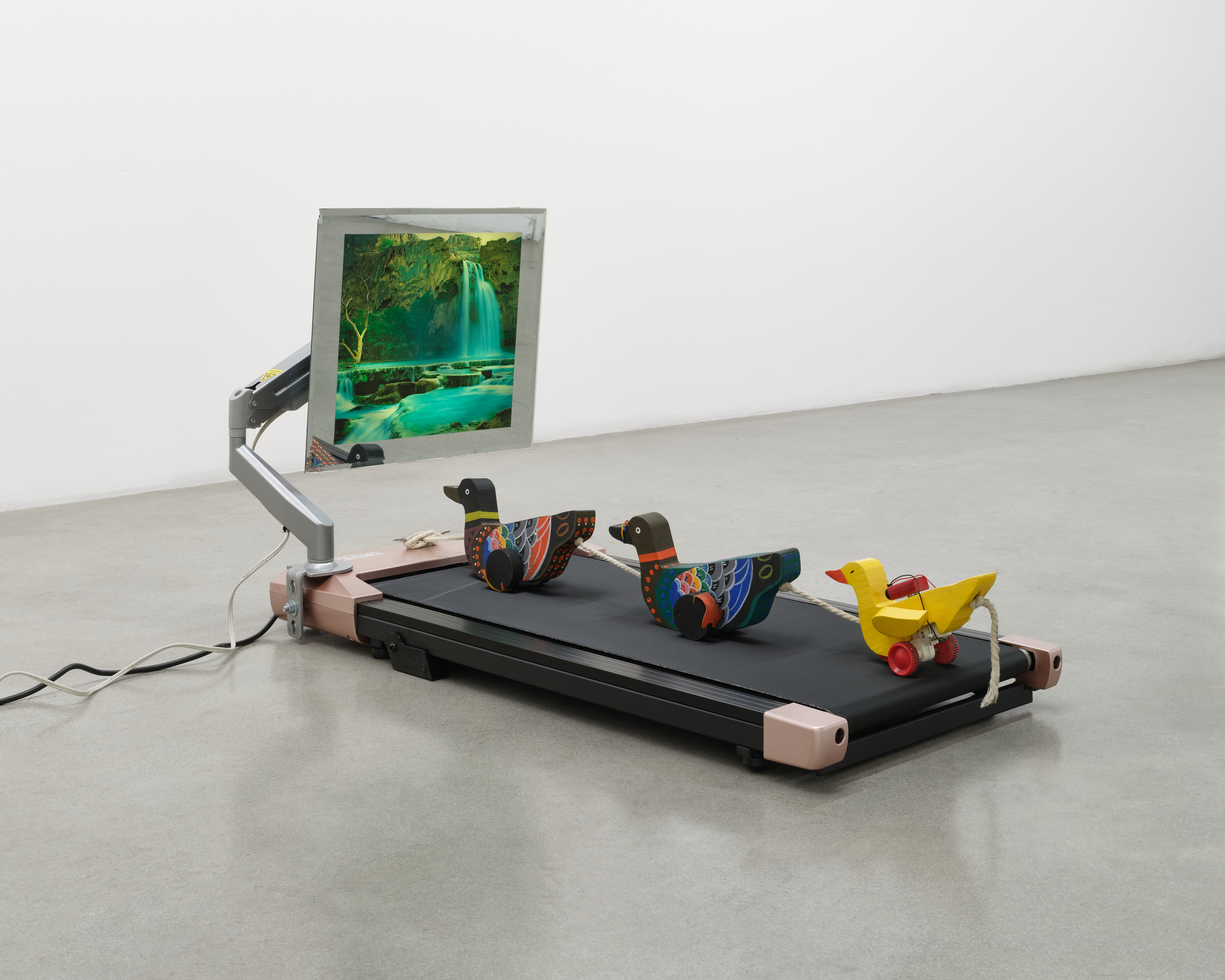 A sculpture by Rachel Youn of three wooden ducks on wheels linked together with rope on a treadmill with an animated photo of a waterfall.