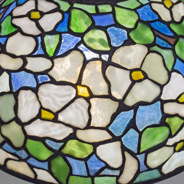 A detail shot of an authentic tiffany lamp shade, the leaded glass design depicting white four-petaled dogwood flowers in mottled and rippled variegated white tiffany glass, with green leaves, against a background of translucent deep blue glass representing the sky
