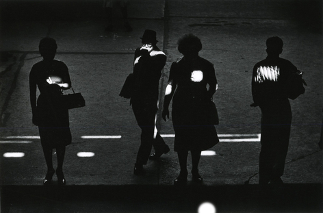 Black and white photo of pedestrians in shadow, with spots of daylight falling on them.