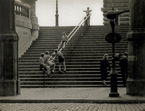 Black and white photo of a group of boys watching each other slide down a railing on a public staircase.