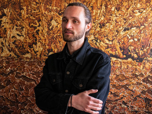 A thin white man cropped at the waist, hair pulled back, wearing a black Demin jacket, stands in front of a painted field of metallic gold and bronze.