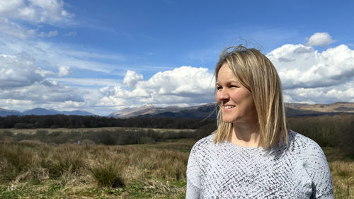 A woman smiling outdoors with a scenic backdrop of rolling hills and a partly cloudy sky.