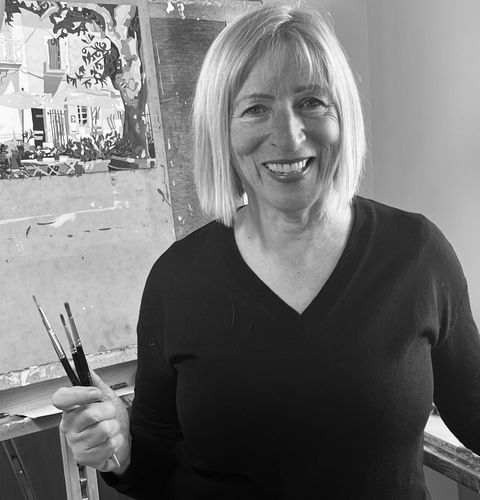 A woman smiling while holding paintbrushes in front of an easel with a partially completed painting.