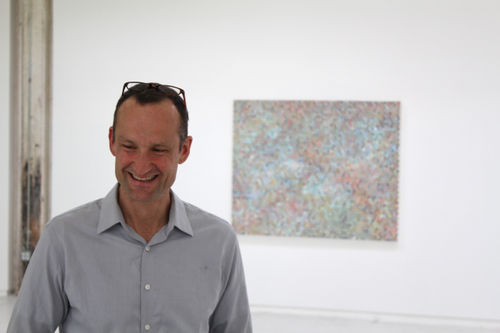 Emil Lukas standing in the gallery with small painting on wall behind him.