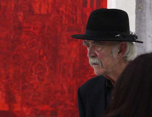 William T. Wiley in an all black complete with a hat, stands in front of a painting with red detailing.