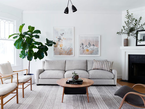 Living room with white and. neutral tones, and green from a few plants. Two works in different sizes by Crystal Liu featuring fantastical landscapes hang above couch.