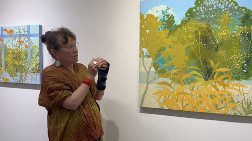 Female artist standing between two abstract landscape paintings hanging on a gallery wall