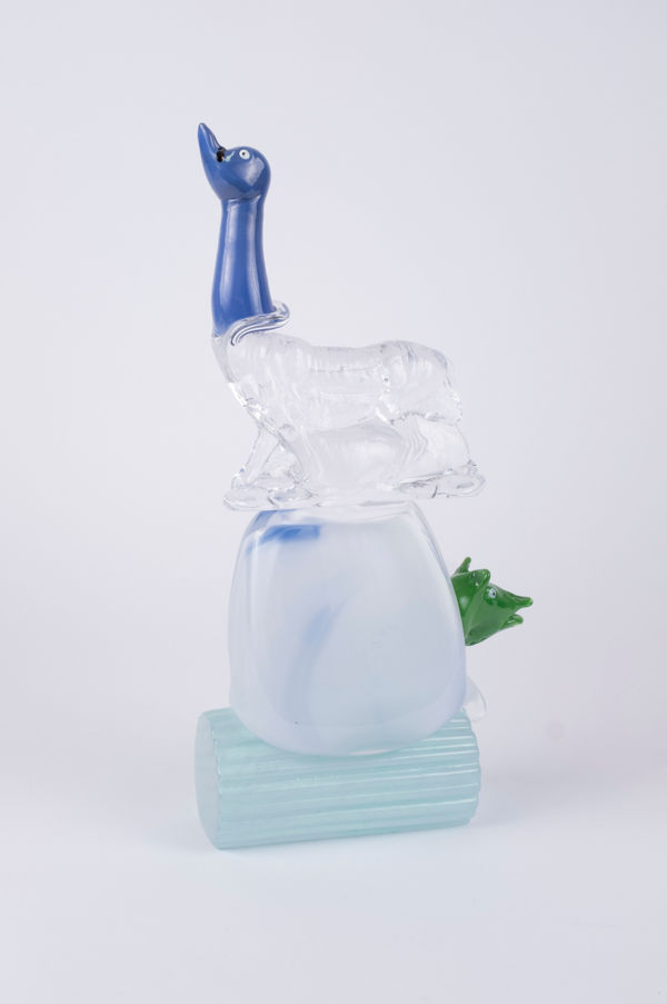 Rasmus Nossbring glasskulptur med ett blått svanhuvud på en vargkropp i klarglas. under en vit kul med ett grönt grishuvud som sticker ut. ovan på en blå liggade pelare.