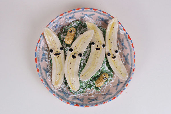 Ceramic sculpture of a bowl of bananas cut in half with faces and two peanuts with faces on a bed of lettuce