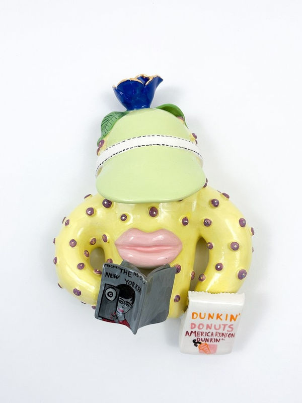 Ceramic creature with large lips holding a book and a Dunkin Donuts bag and wearing a green visor