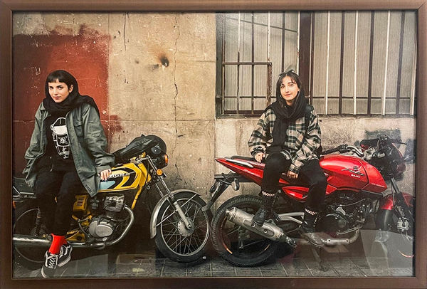 Photo of two women sitting on red and yellow motorcycles in wooden frame