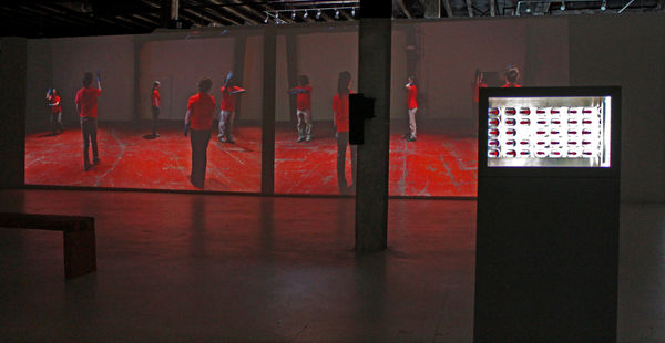 Still from large video installation. In video, two people are wearing red or blue polo shirts, black pants and blue latex gloves. They are dancing in a stiff, mechanical fashion.