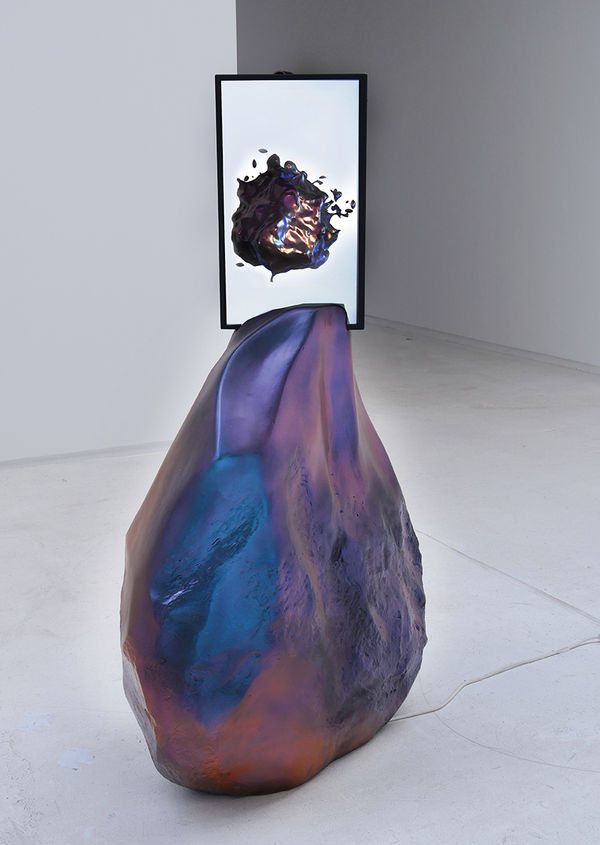 Chrome colored boulder with two screens mounted on top. The screens show stills from the videos of a fluid chrome shape metamorphosing into other shapes.