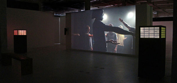Still from video installation. In video, people standing on scratched red floor is are dancing in a stiff and mechanical manner.