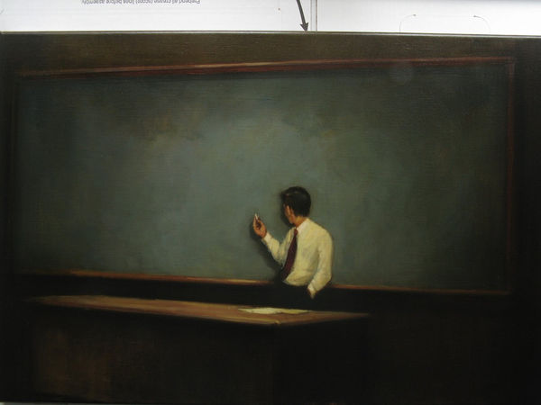 Man wearing tie stands in front of chalkboard holding a piece of chalk up with one hand and with other hand in his pocket. His face is hidden from the viewer as he looks towards the chalkboard.