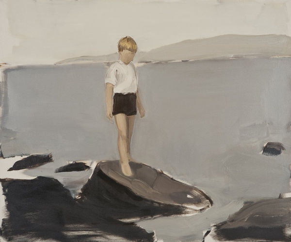 Young faceless blonde boy in white shirt and black shorts stands on rocks at the shore of a lake with water, mountains and sky in the background.