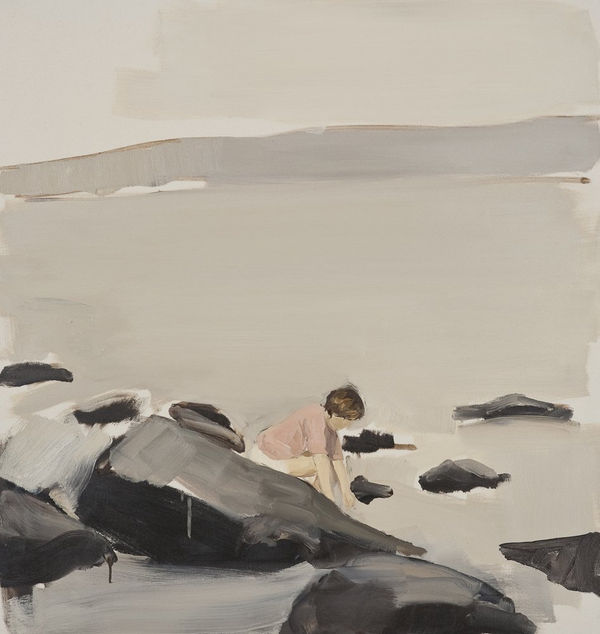 Child is hunched over seated on rocks at a lake’s shore with gray water, mountains and sky in the background.