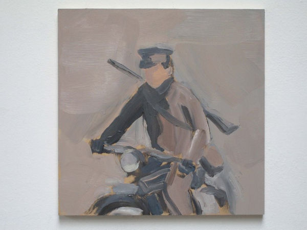 Faceless soldier in uniform rides motorcycle with guns strapped to his back in front of gray background.