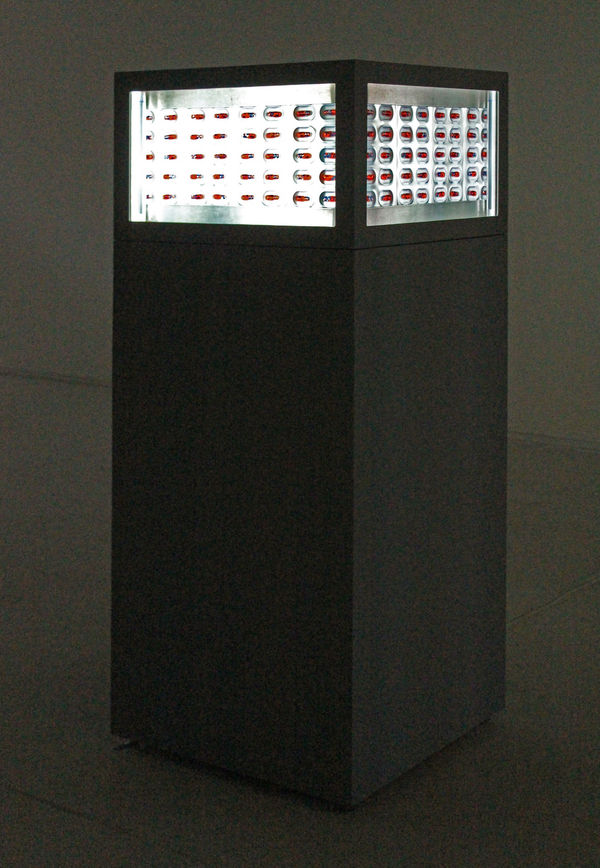 Still from video installation. Tall black box with four screens mounted near the top of each side. The screens show rows of red pills pointed horizontally.