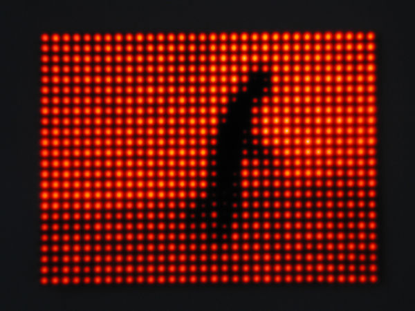 Still image of a rectangular piece with red LEDs behind Plexiglass that displays a pixelated shadowy man walking