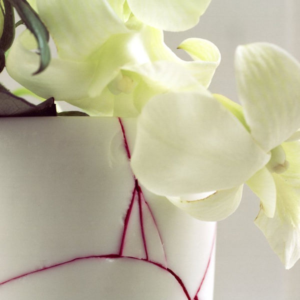 Digital c-print of a photograph which depicts a zoomed in view of two green white orchid blossoms handing down and out of a white ceramic vase. The vase has several cracks filled with deep pink ink