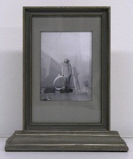 Black and white scene of person wearing shapeless jumpsuit and standing in room filled with assortment of shapes and objects.