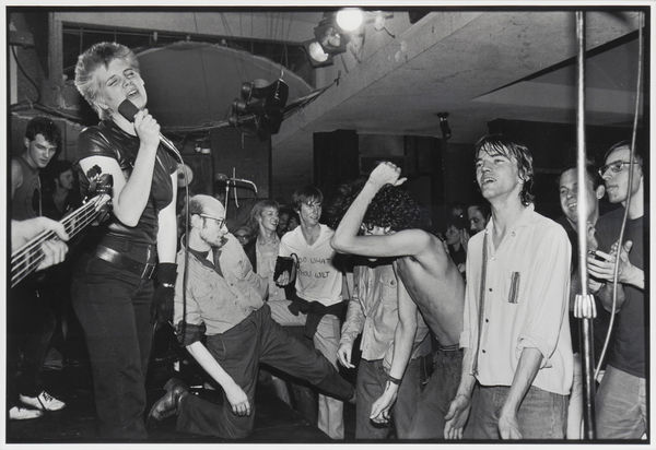 Black and white photograph of punk rock group The Avengers performing at a concert In the scene lead singer Penelope Houston sings on stage on the stage while surrounded by sound and light equipment along with rowdy members of the audience