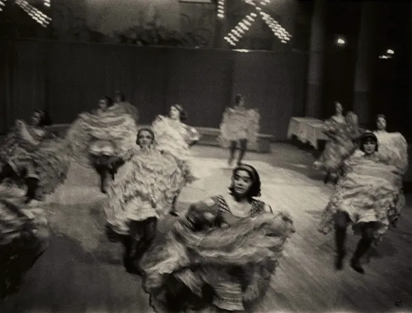 Ilse Bing photograph of chorus dancers performing on a stage, their ruffled skirts swirling in motion as they move toward the camera under theatrical lights, black-and-white.