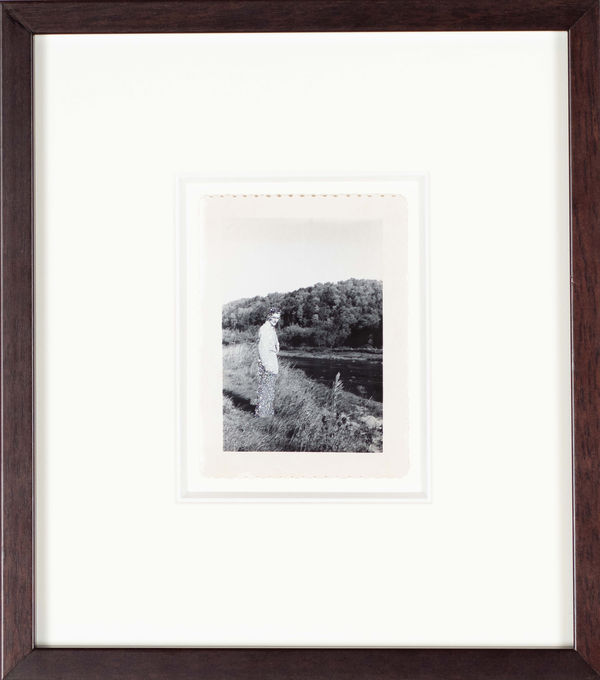 John Latour, Man in suit standing near the river’s edge with head turned towards the camera, 2015