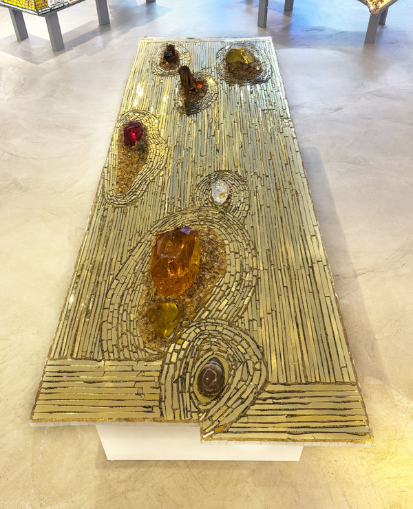 A rectangular gold sculpture by artist Andrew Logan with circular patterns like a Zen garden. It has small glass pieces and stones embedded in its surface that reflect light.