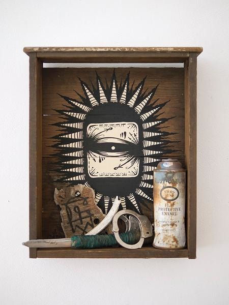 Wooden shelf with painting of an eye by GATS with several trinkets in the shelf including a spray can, handcuffs, piece of wood and a knife
