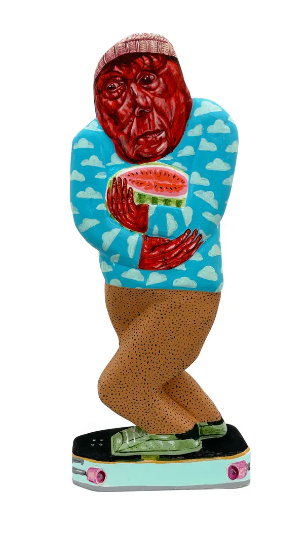 Sculpture of a man riding a skateboard holding a cut open watermelon. There are two different but similar sides. This side of his face are red and he is wearing a blue sweater with light blue clouds on it. There is a red beanie on his head.