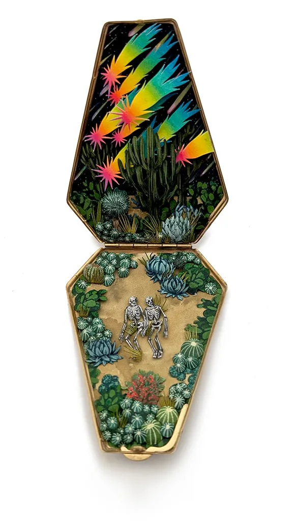 hand-cut watercolor collage in antique cosmetic compact by Shannon Taylor of two skeletons laying down on the ground with cactus surrounding them and colorful stars above them