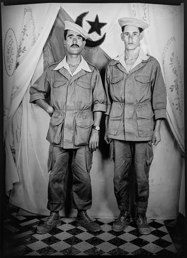 1960s black and white photograph of two men standing wearing berets and a military khaki uniform in front of the Algerian flag