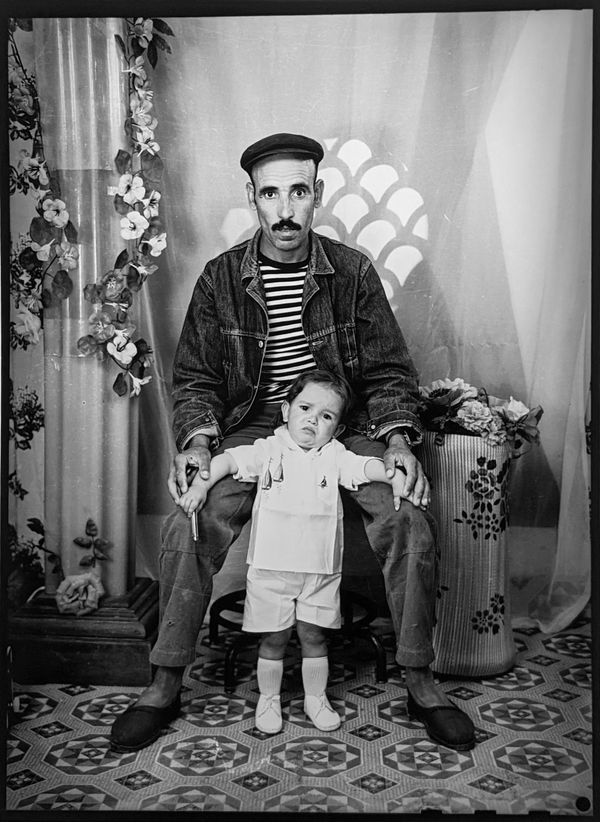 1960s black & white photograph of a seated man in striped T-shirt holding the hands of a young boy standing, in a photo studio