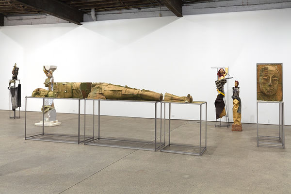 Installation view of Matthew Monahan's sixth solo show at Anton Kern Gallery, featuring sixteen sculptures, a set of drawings, two wall-mounted masks, and a single large figure in the back room. Long sculpture with other works.