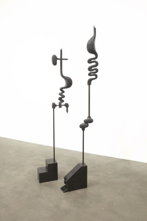 Two thin vertical Eric Fertman sculptures that appear to be wearing shoes