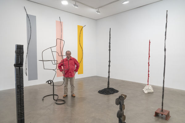 Installation View of Susan Inglett Gallery featuring Alain Kirili with several of his vertical sculptures and strips of gray, peach, and yellow on the wall behind him