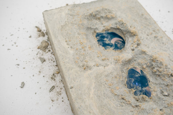 A sculpture by Shabez Jamal consisting of a photograph of an older woman in glasses and a pink hat. The photograph is submerged in a block of concrete but partially visible.