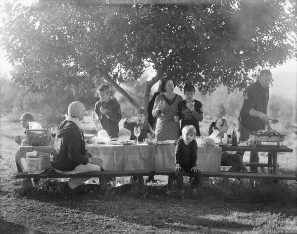B&W family portrait by Ariel Yannay. A large group gathers around a table outdoors, documented in soft, large-format light to explore the presence of those who follow us.