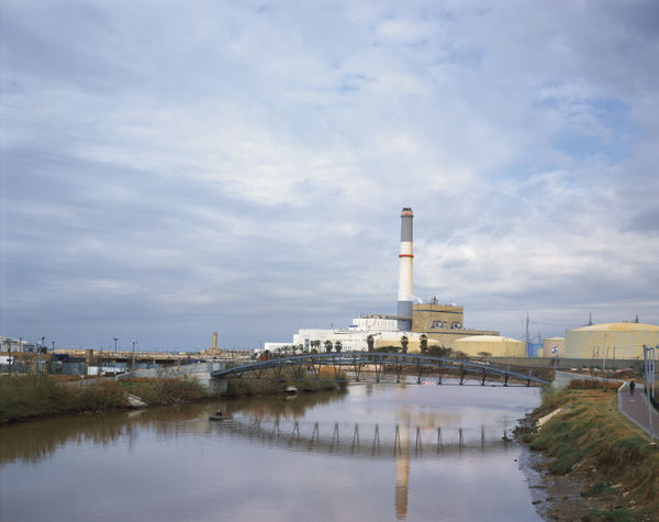 A view of the Yarkon River leading toward the Reading Power Station chimney and a modern arched bridge.