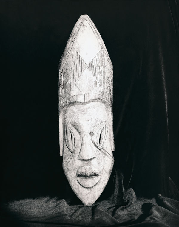 A high-contrast print of a carved mask, showing the sharp vertical wood grain against a void of pure black.