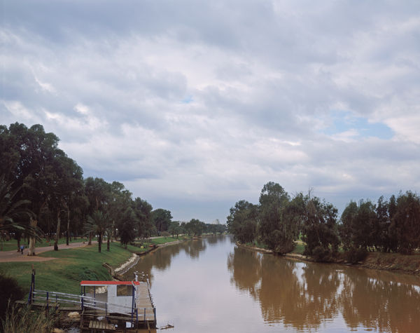 A view of the Yarkon River with a small white pier in the foreground and lush trees lining the banks under a cloudy sky.